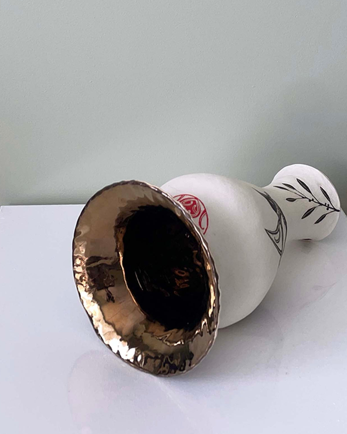 A white ceramic vase with a metallic gold interior shown lying on its side on a white surface against a light gray wall. The vase has a small red floral accent and a black leaf pattern on the neck.