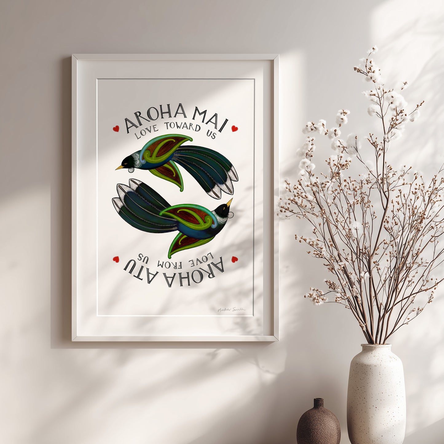 Framed artwork with birds and 'Aroha Mai' text on a wall with a vase of dried flowers.
