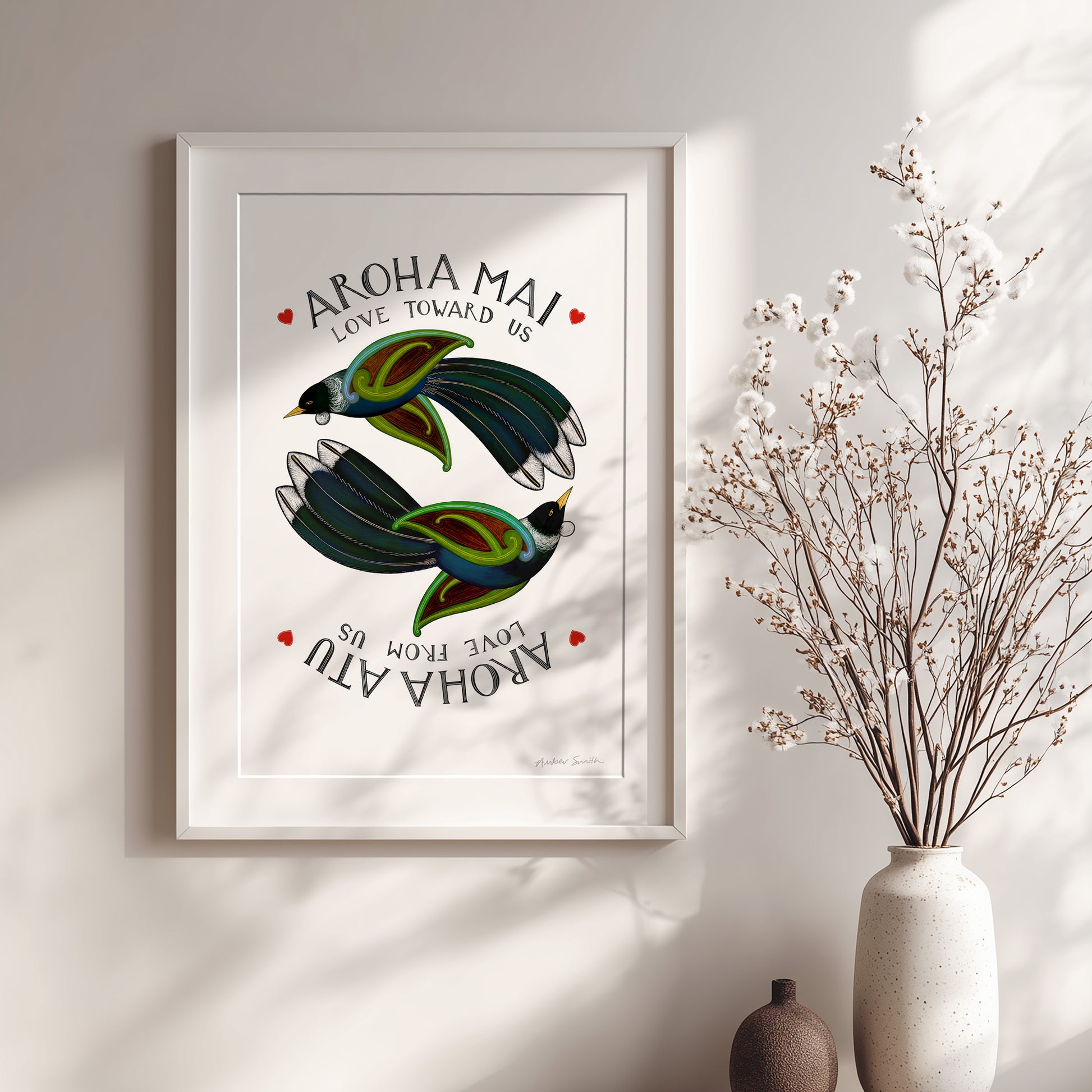 Framed artwork with birds and 'Aroha Mai' text on a wall with a vase of dried flowers.