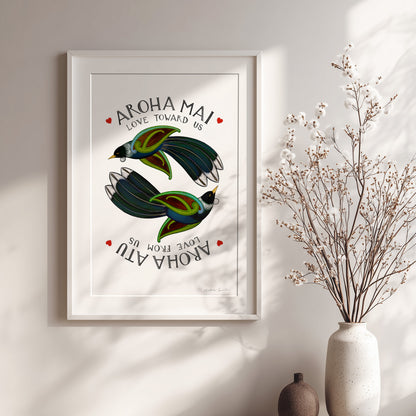 Framed artwork with birds and 'Aroha Mai' text on a wall with a vase of dried flowers.