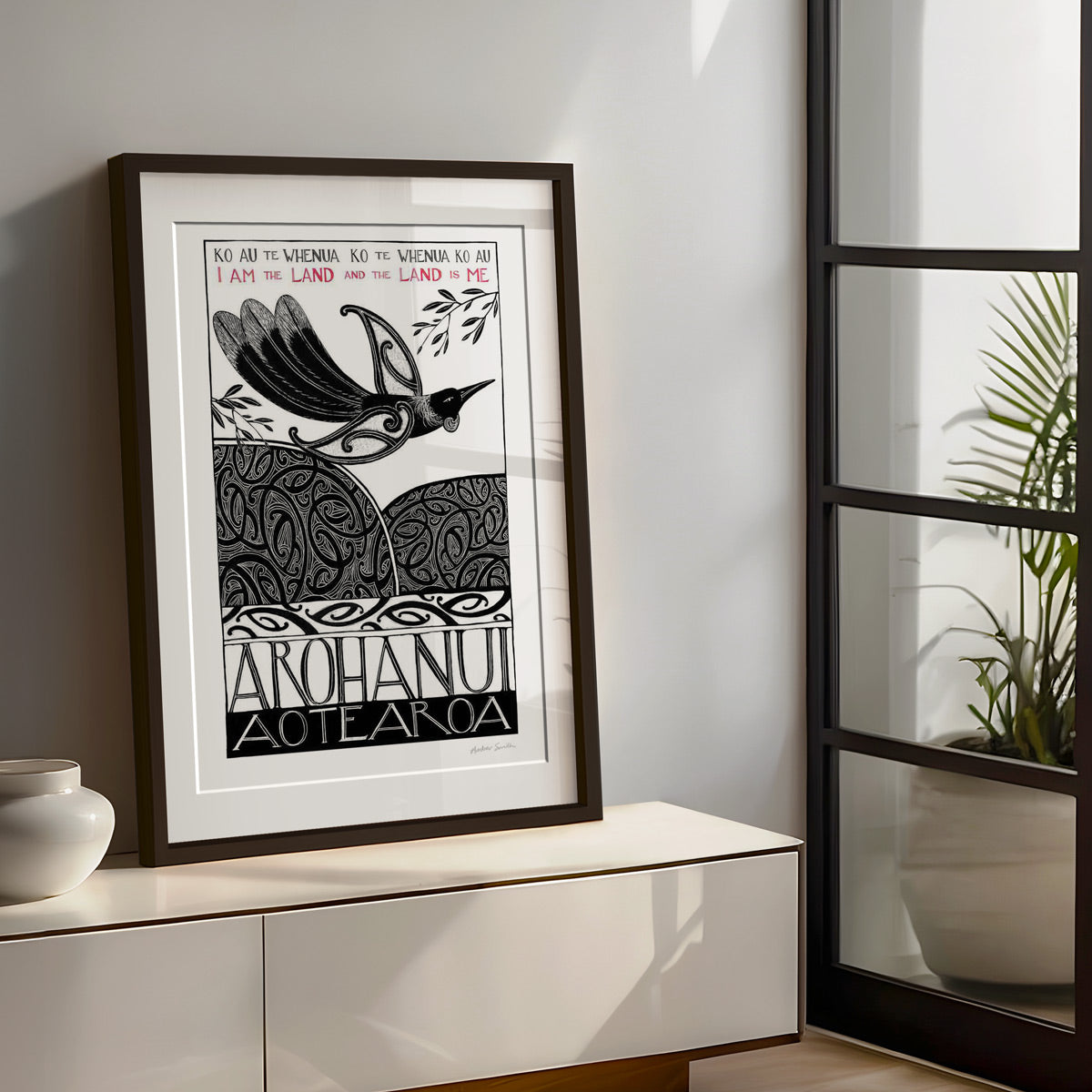 Large framed New Zealand tūī art print, Ko au te Whenua – I Am the Land, featuring a tūī flying over the land and sea, with the te reo Māori whakataukī “Ko au te whenua, ko te whenua ko au” and its English translation, “I am the land and the land is me”.I am the land and the land is me te reo whakatauki art print with tui bird flying over koru pattern hils and sea
