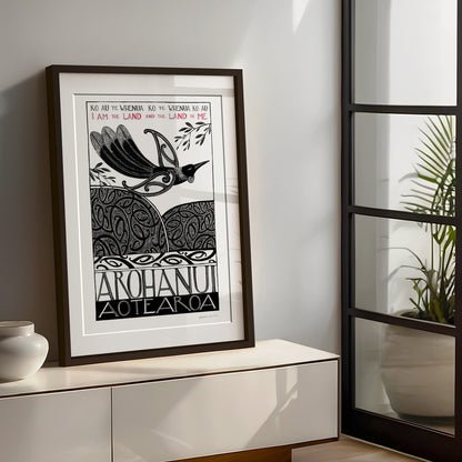 Large framed New Zealand tūī art print, Ko au te Whenua – I Am the Land, featuring a tūī flying over the land and sea, with the te reo Māori whakataukī “Ko au te whenua, ko te whenua ko au” and its English translation, “I am the land and the land is me”.I am the land and the land is me te reo whakatauki art print with tui bird flying over koru pattern hils and sea
