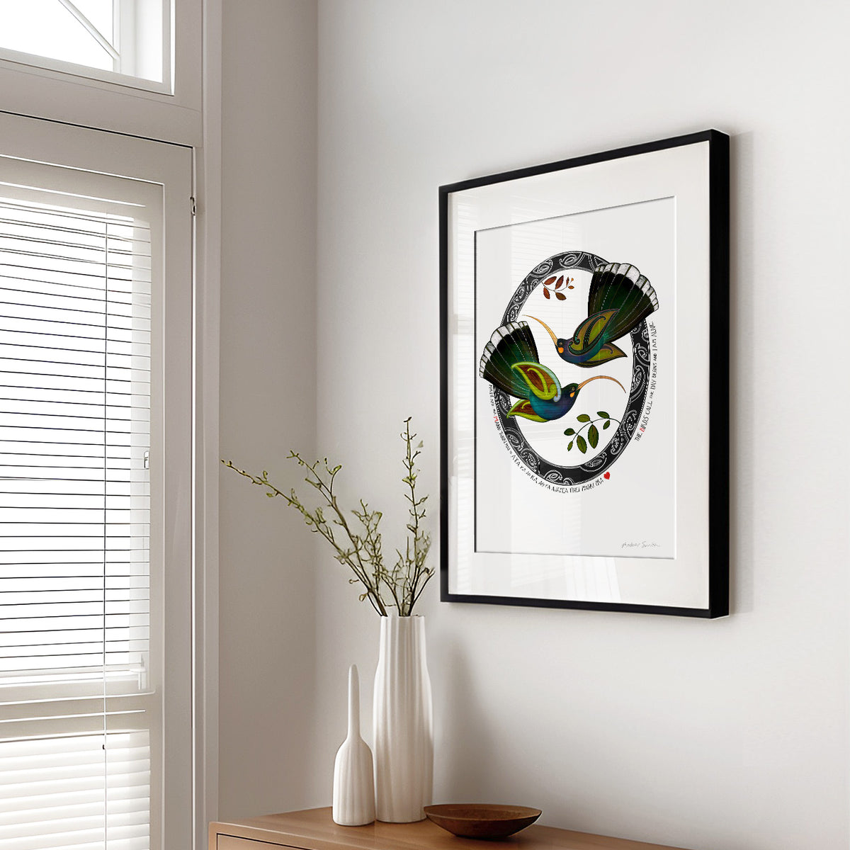 New Zealand huia bird art print, Tūmanako – Hope, featuring two huia taking flight into the light of a new dawn, with a te reo Māori karakia about the birds calling and the day beginning. Shown in living room with black frame.